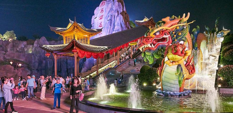 Colorful dragon statue and pavilion at a night festival, surrounded by people and fountains.