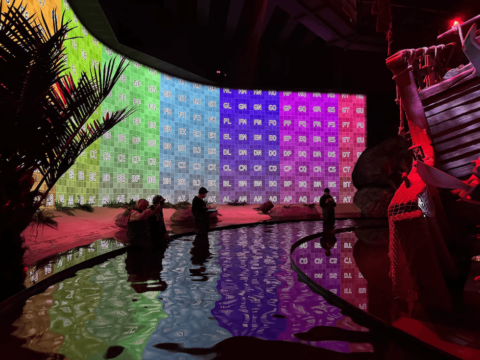 Colorful LED screen wall with reflections and silhouettes of people near water in an indoor setting.