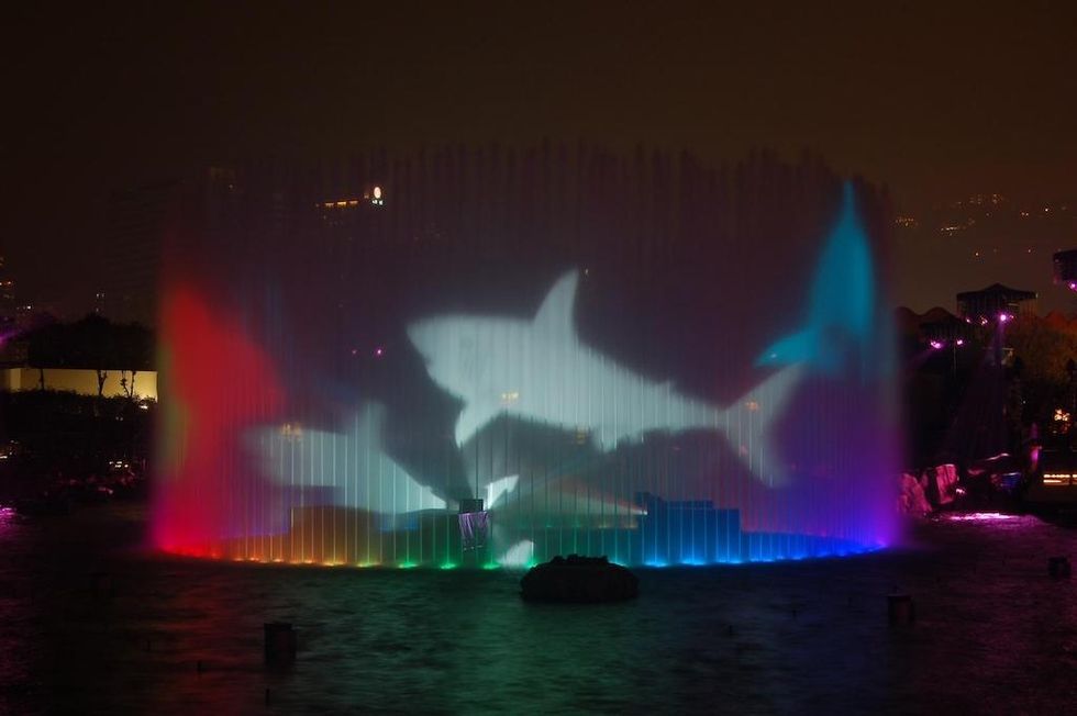 Colorful light show with shark projections on a water fountain at night.