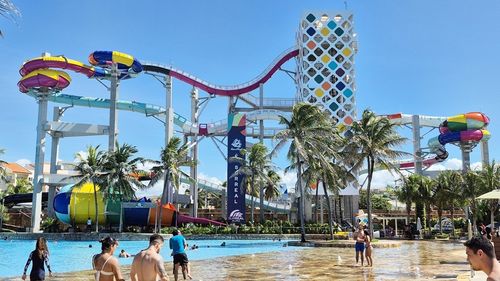 Colorful water park with winding slides, palm trees, and people enjoying a sunny day in the pool.