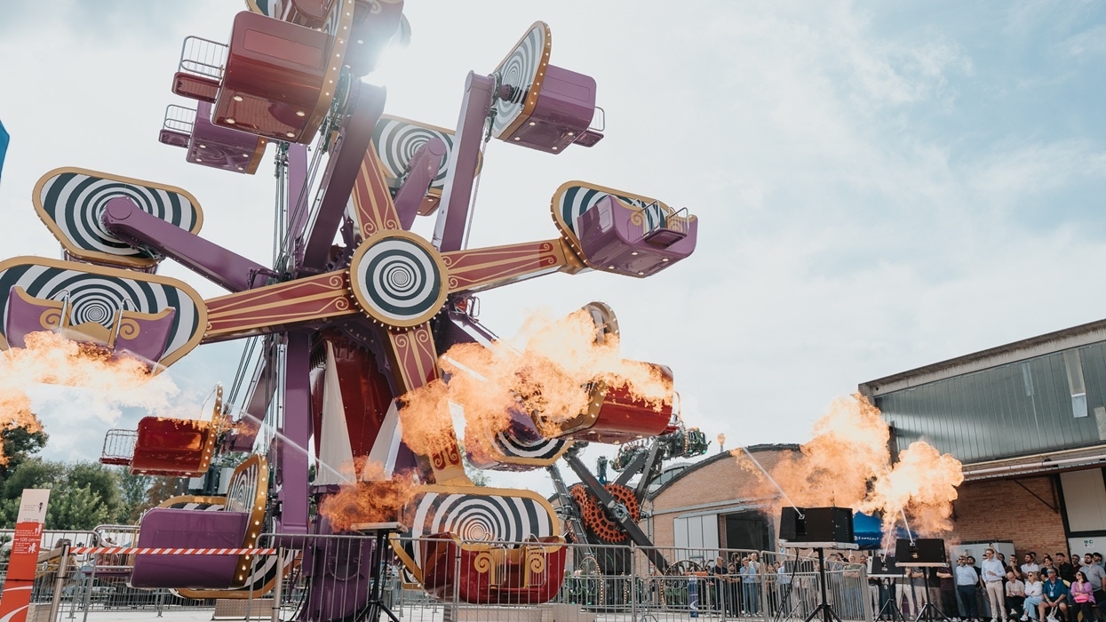 Colourful Hypnosiz ride with spinning arms, surrounded by fire effects, and a crowd watching.