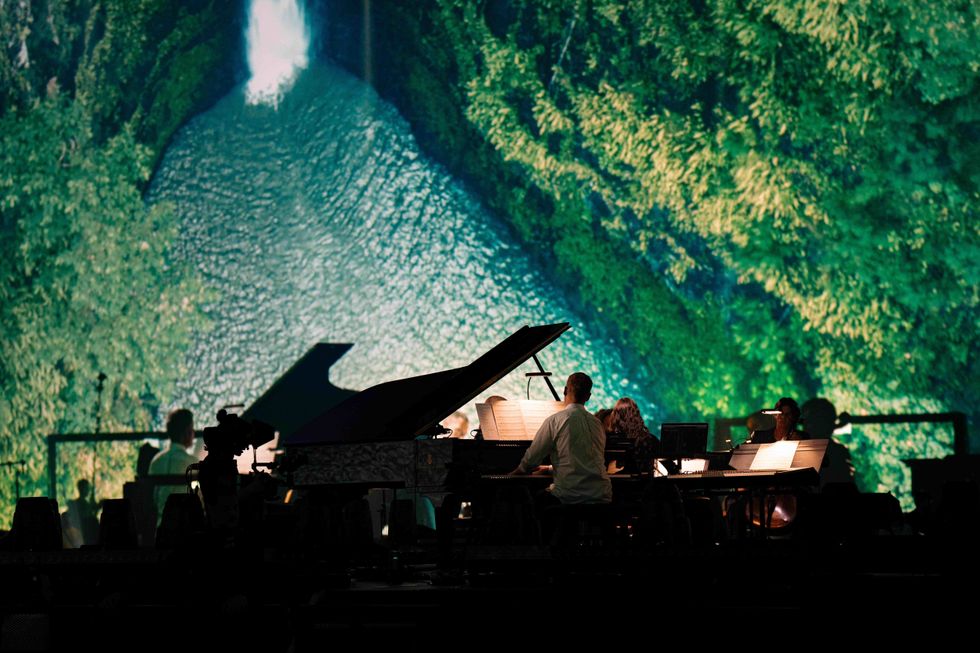 Concert pianist performs on stage against a vivid waterfall backdrop.