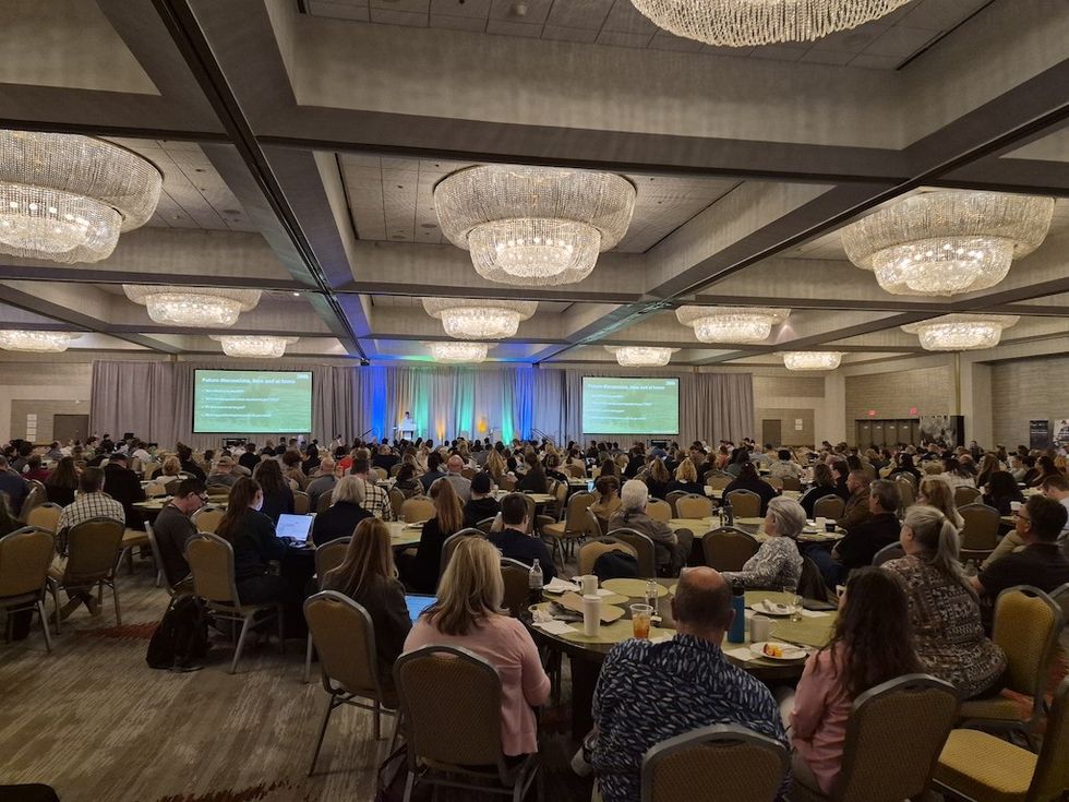 Conference attendees seated at tables, facing a stage with presentations on large screens.