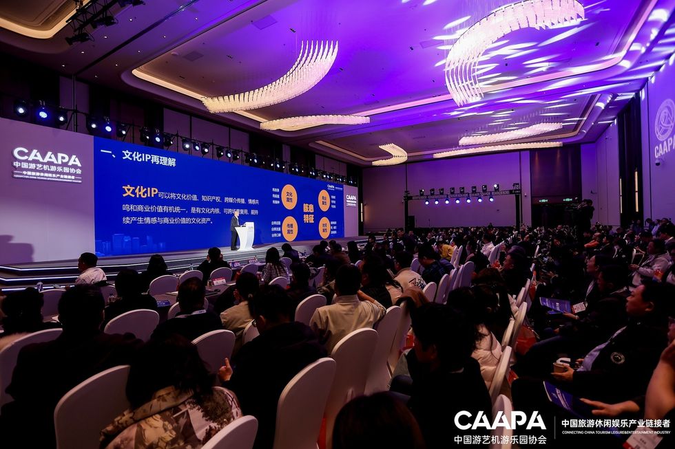 Conference audience seated facing a stage with a presentation display.