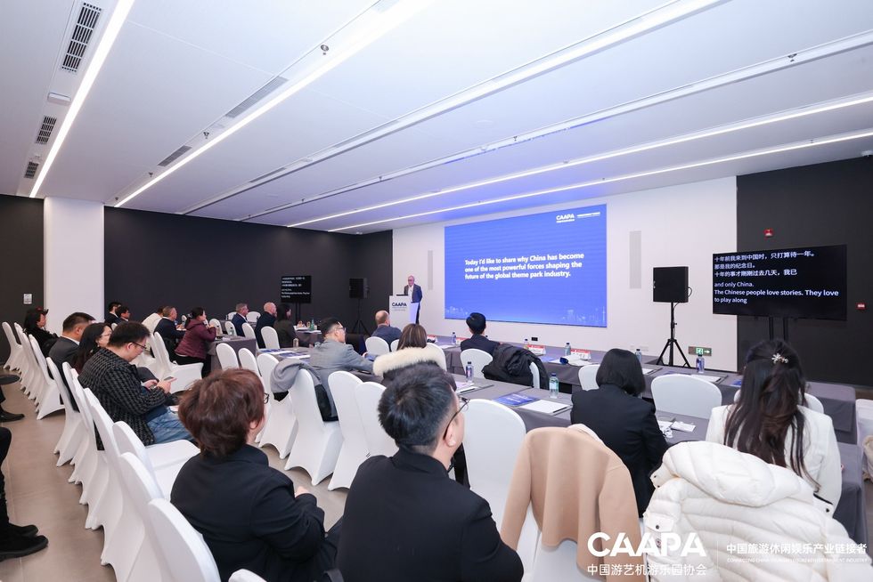 Conference room with a speaker presenting to an audience; large screen displays text.