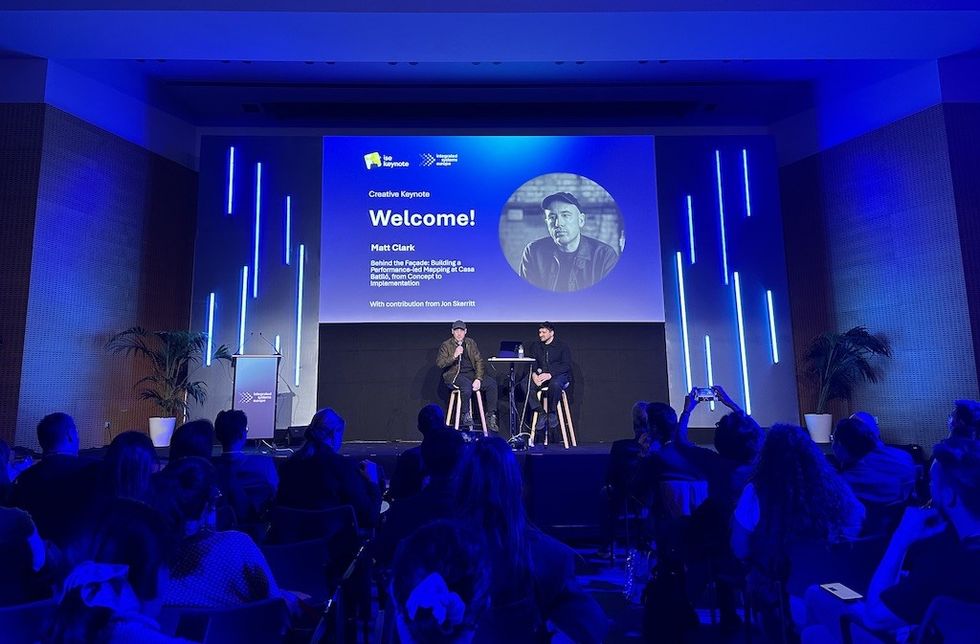 Conference talk with two speakers on stage, large screen displaying welcome message.