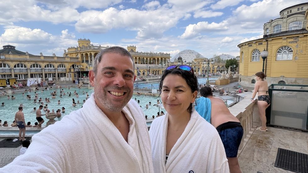 Couple in robes at Sz\u00e9chenyi Thermal Bath, Budapest, with crowded pool and historic building.