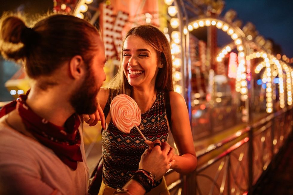 Couple laughing at a fairground with a large lollipop and bright lights in the background.