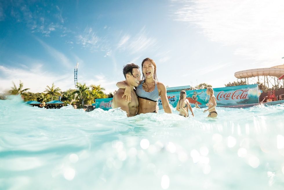 Couple laughing in a sunny water park pool, surrounded by others and palm trees.