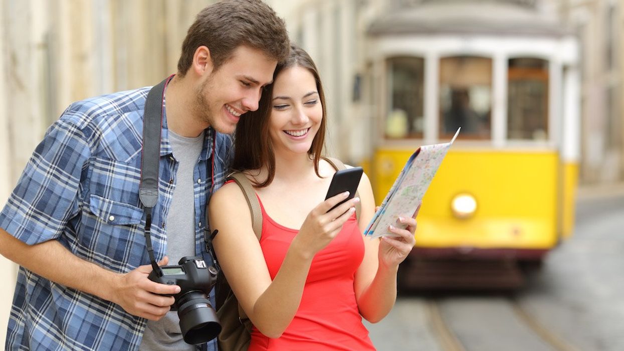 Couple with camera and map, smiling while checking phone, with a yellow tram in the background.