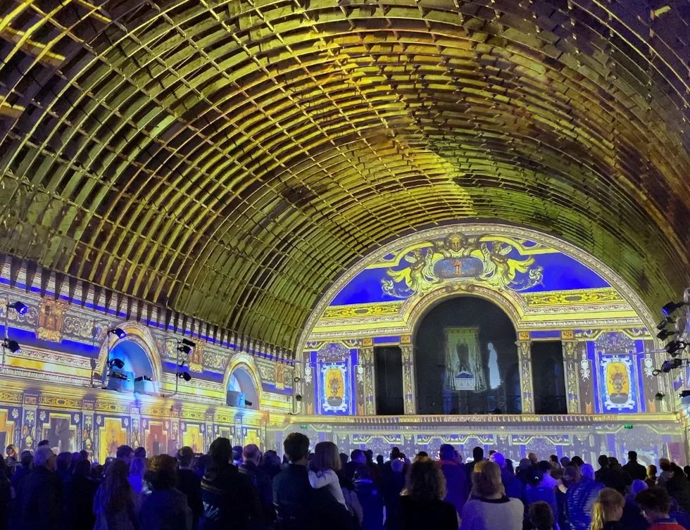 Crowd viewing colorful projections on ornate arched interior with vaulted ceiling.