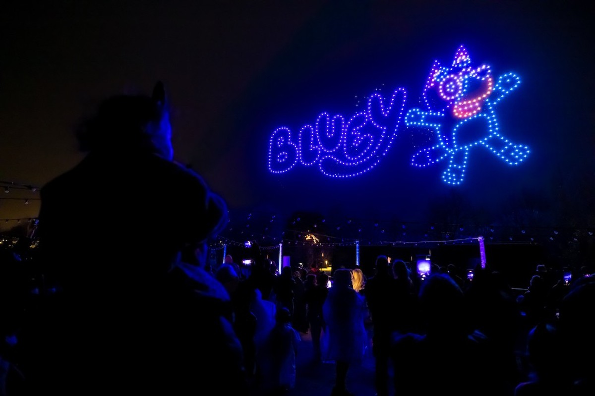 Crowd watches a colorful drone display spelling "BLUEY" with character in the night sky.