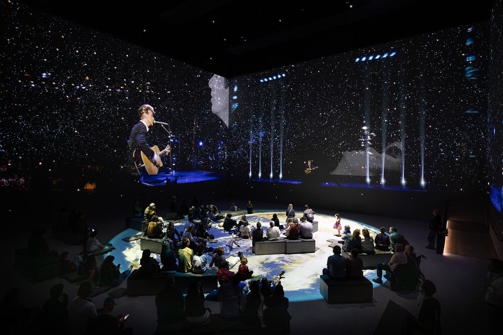 Crowd watching a large projection of a guitarist under a starry night sky in a dark room.