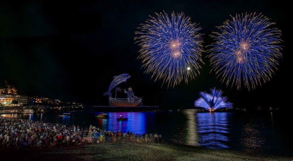 Crowd watching blue fireworks and drone light show over a beach at night.