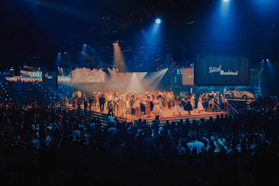 Crowded concert hall with performers on a lit stage under blue and orange lights.