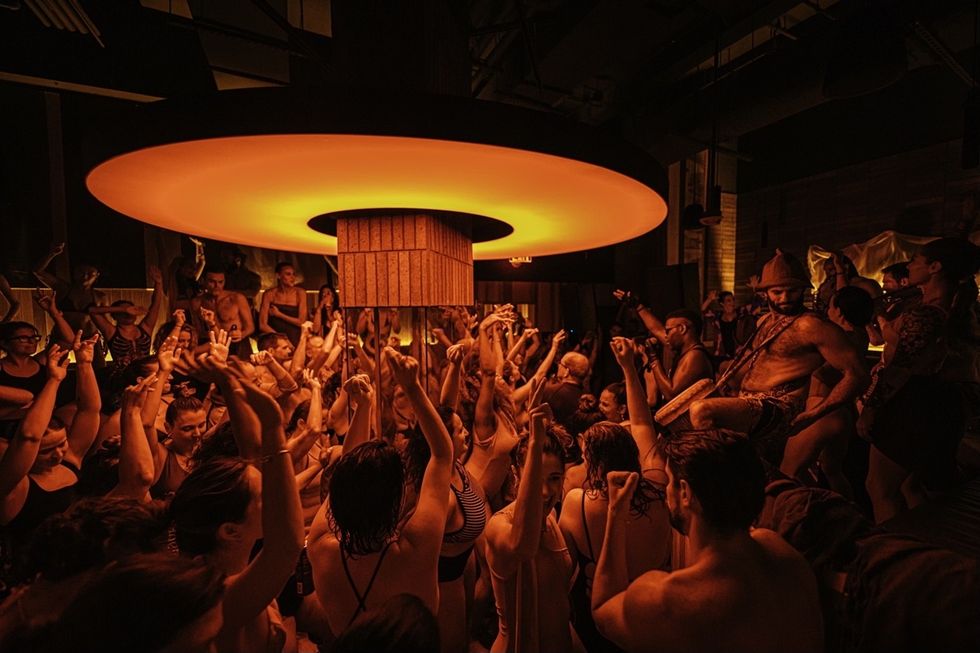 Crowded dance floor with warm orange lighting and people raising their hands in enjoyment.