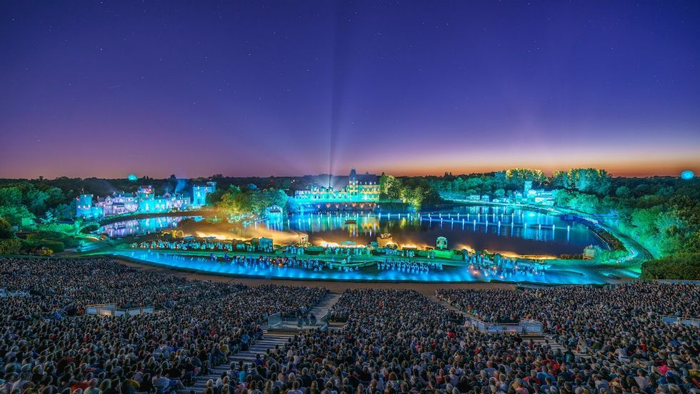Crowded outdoor night showm \u200bLa Cinesce\u0301nie at Puy du Fou, with illuminated historic buildings and water reflections.