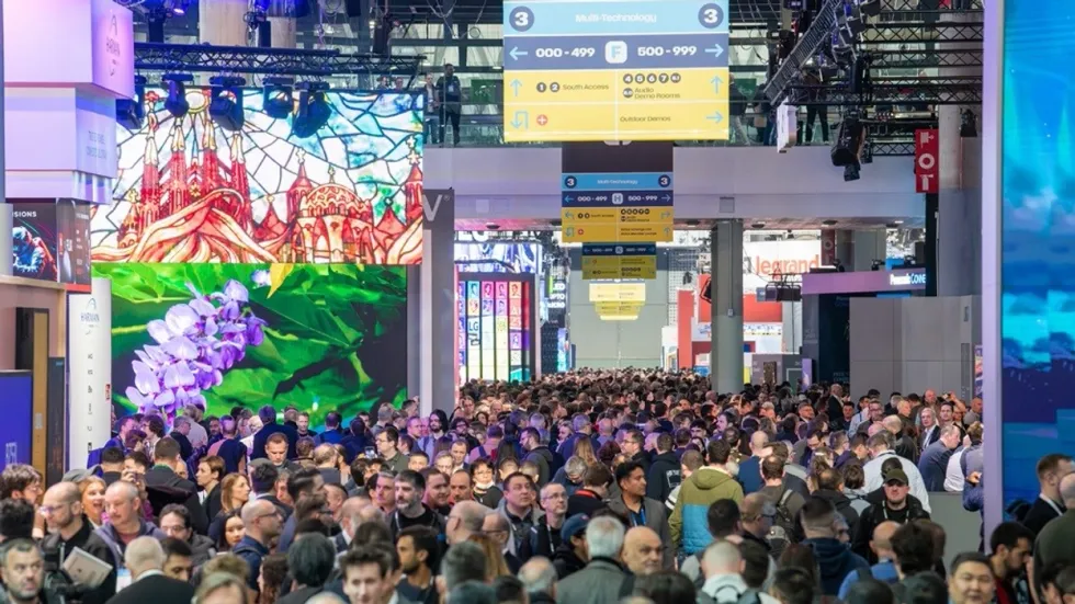 Crowded technology expo hall with vibrant displays and directional signs above - ISE