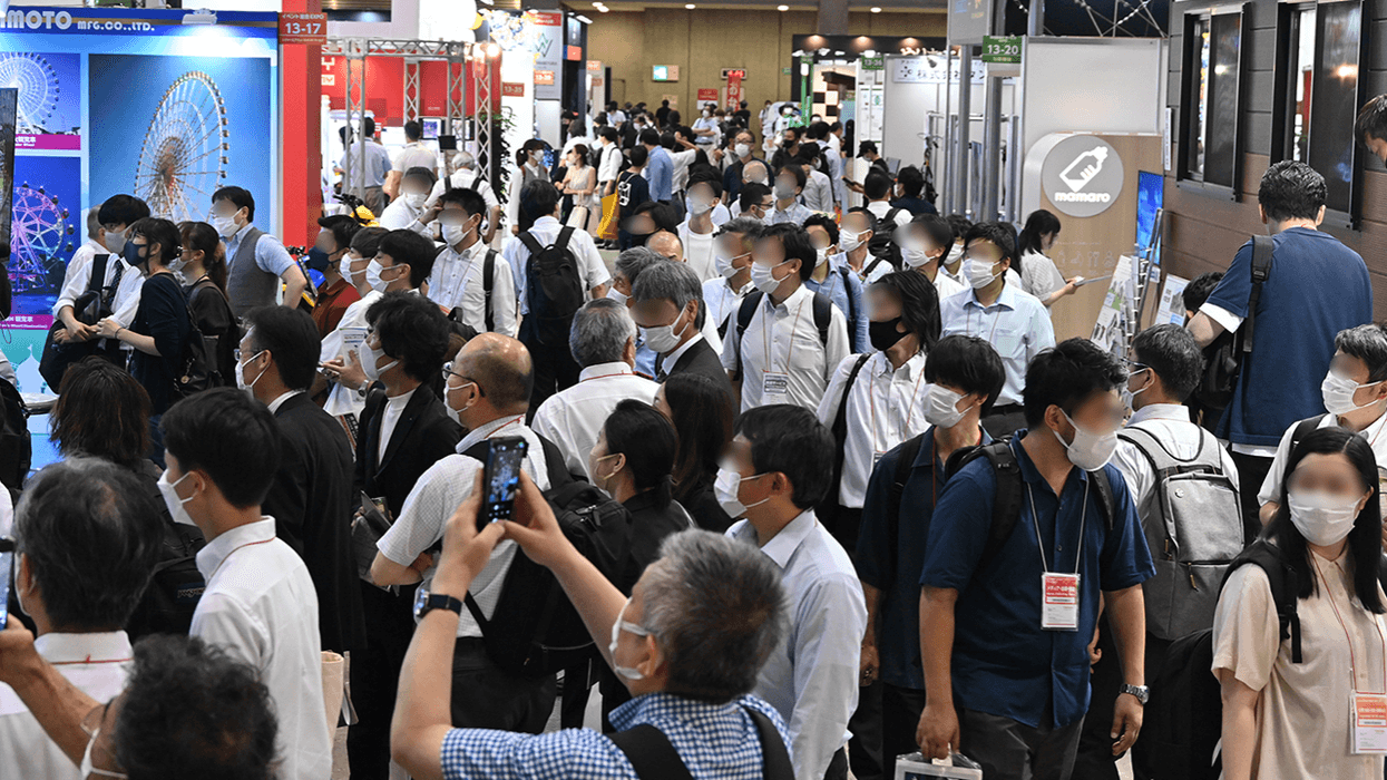 Crowded trade show with masked attendees exploring booths and displays.