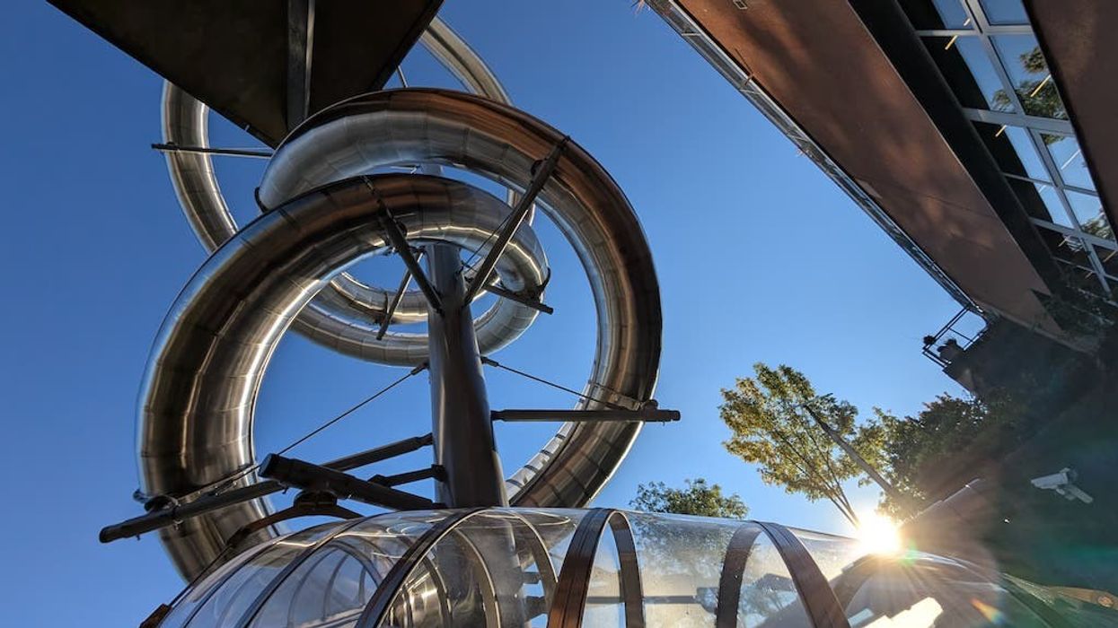 Curved metallic slide from Wiegand at Moderna Museet, against a clear blue sky with sunlight peeking through.