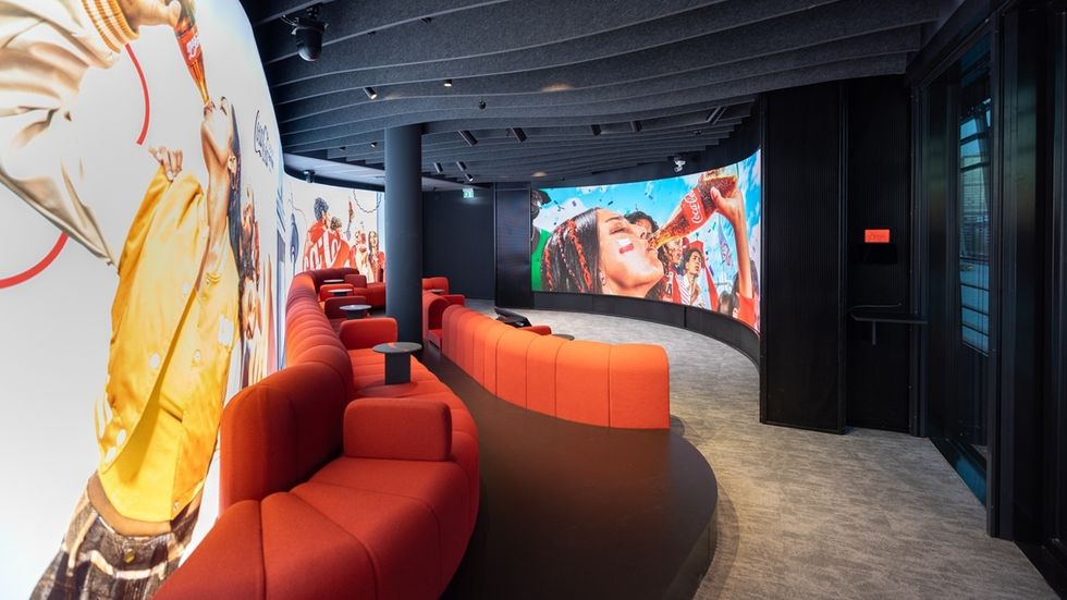 Curved room with red chairs, Coca-Cola murals, and a large screen displaying happy people at THE 6TH FLOOR customer experience centre.