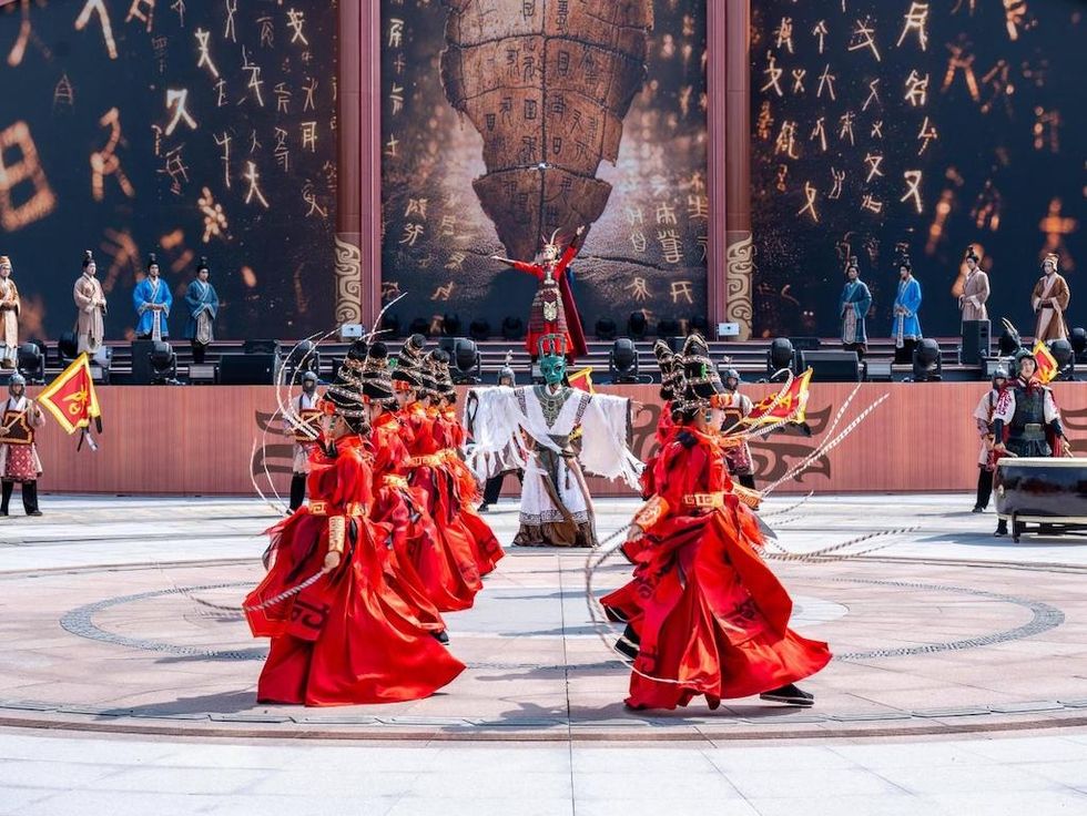 Dancers in red costumes perform on stage with a dramatic background of ancient symbols.