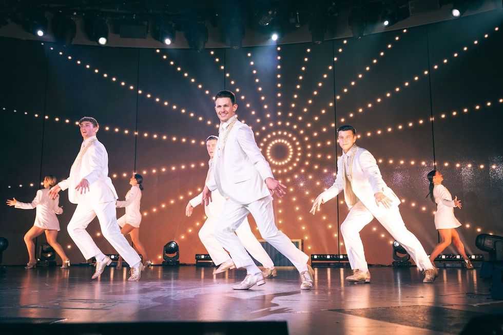 Dancers in white performing on stage with a radiant light backdrop.