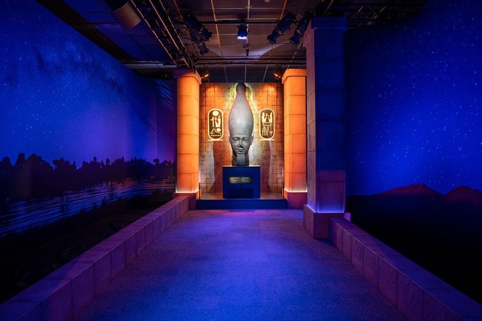 Dimly lit room with a large ancient Egyptian statue and hieroglyphs on a vibrant, blue backdrop.