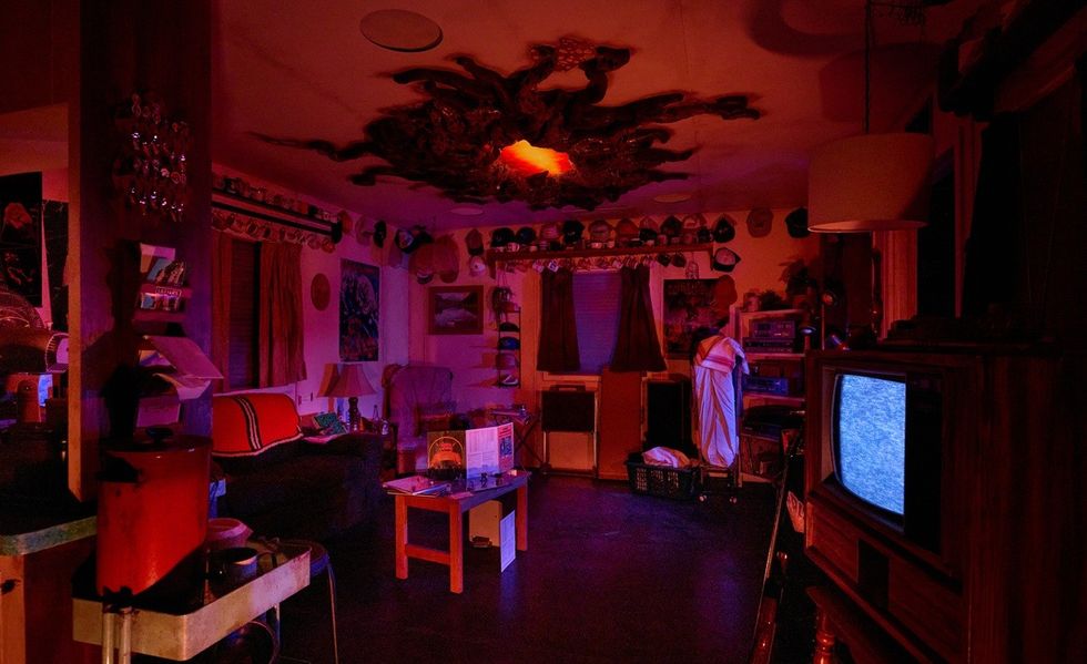 Dimly lit room with red lighting, vintage TV showing static, and cluttered decor, with glowing orange rift in ceiling. Stranger Things: Face the Dark Experience at Netflix House Dallas