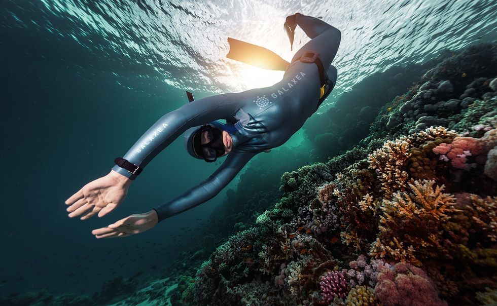 Diver glides over vibrant coral reef in clear ocean water, sunlight in the background.