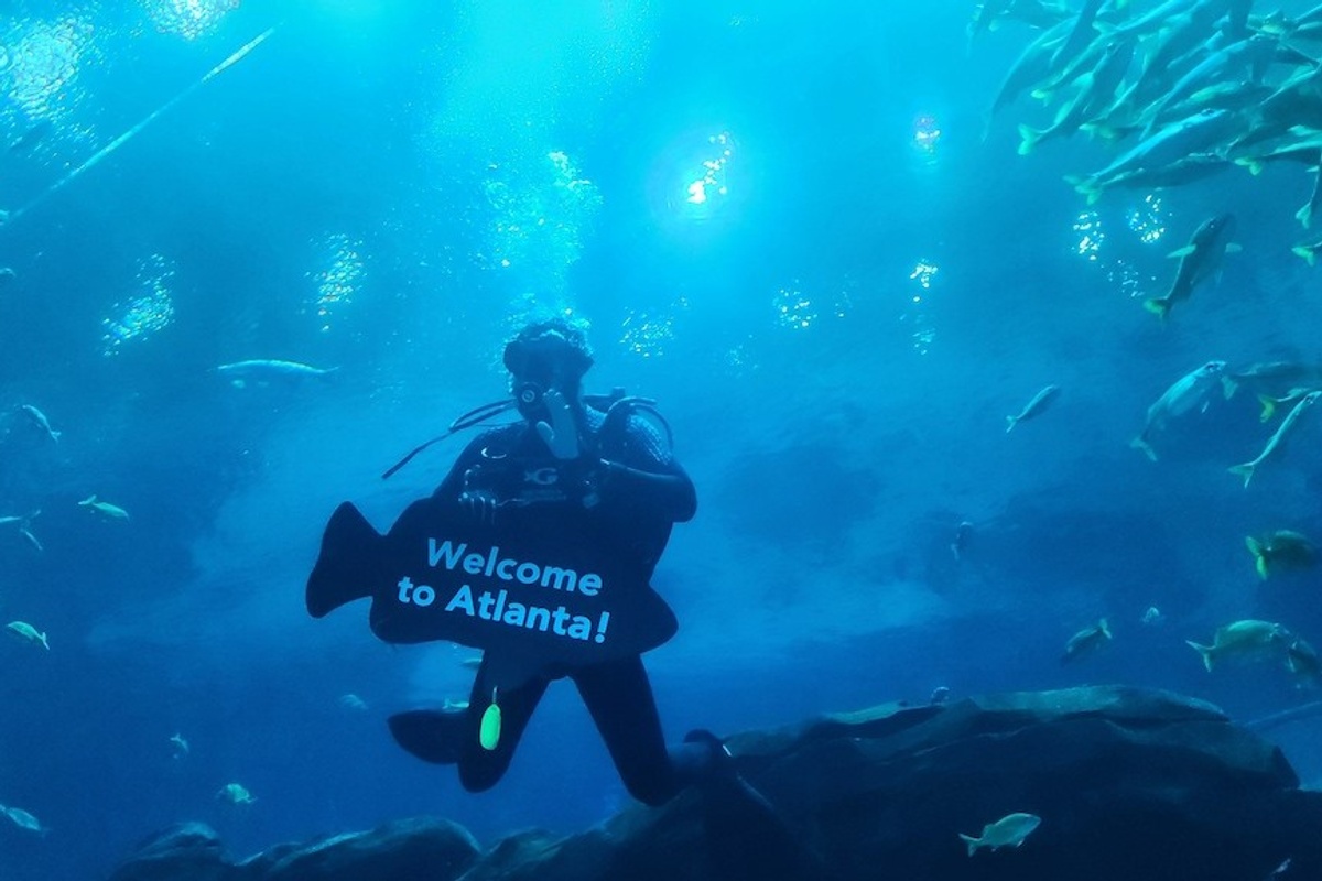 Diver holding "Welcome to Atlanta!" sign surrounded by fish underwater.