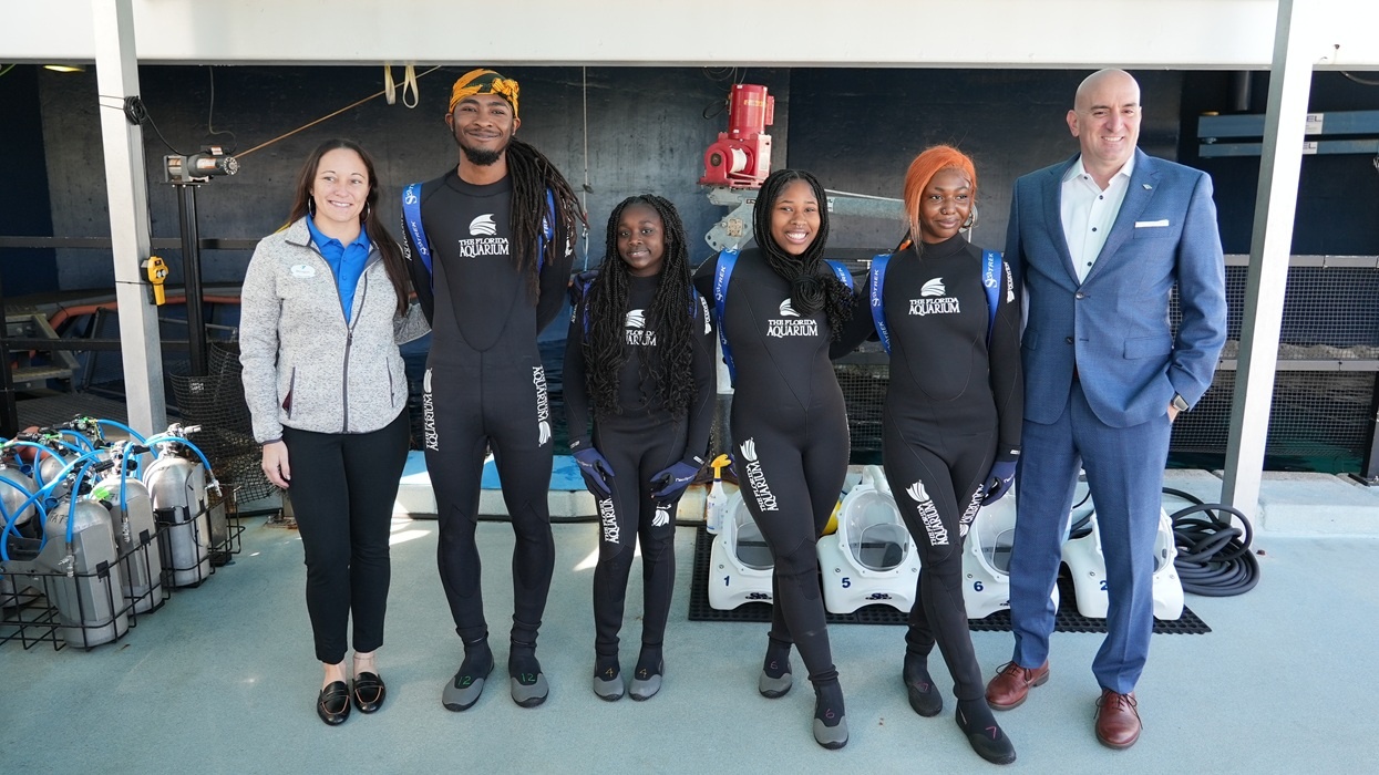 Diverse group in wetsuits poses for a photo at an indoor aquatic facility at the Florida Aquarium's SeaTREK experience