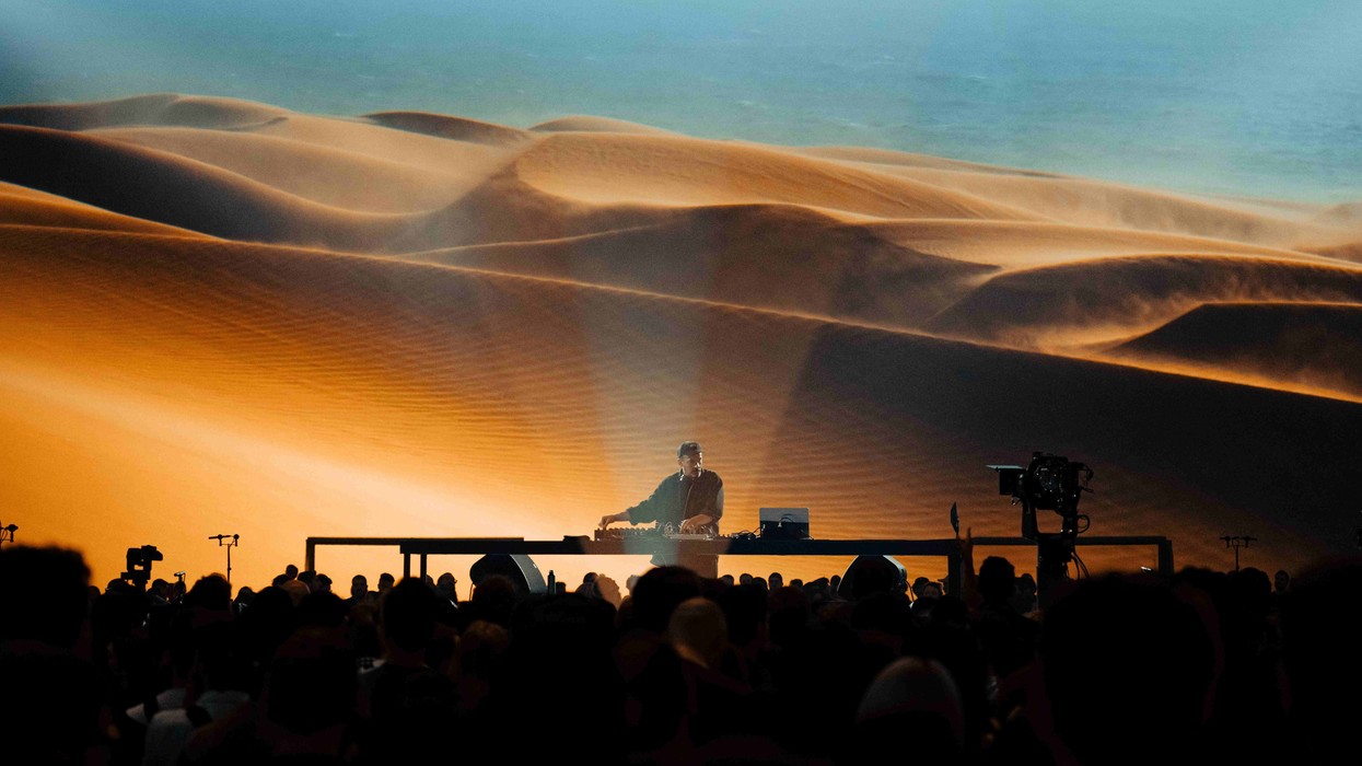 DJ performing in front of a large desert landscape backdrop, with a crowd watching.