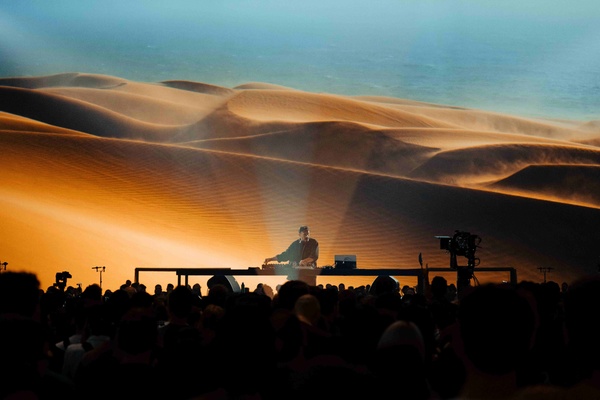 DJ performing in front of a large desert landscape backdrop, with a crowd watching.
