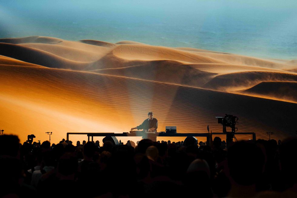 DJ performing in front of a large desert landscape backdrop, with a crowd watching.