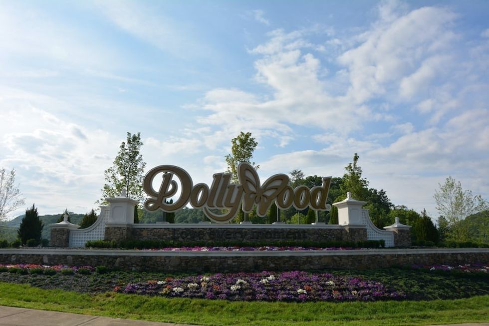 Dollywood entrance sign with butterfly logo, surrounded by flowers and greenery under a blue sky.