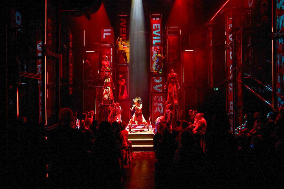 Dramatic red-lit theater scene with performers and illuminated text in the background.