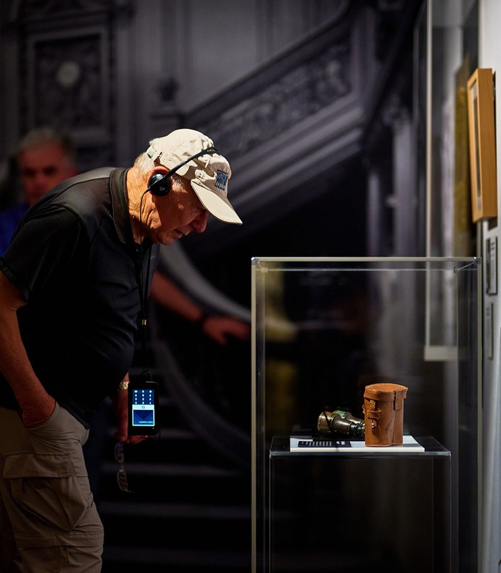 Elderly man examines museum exhibit, wearing headphones and a cap.