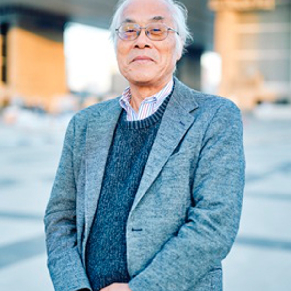 Elderly man in a gray suit stands outside with a modern building in the background.