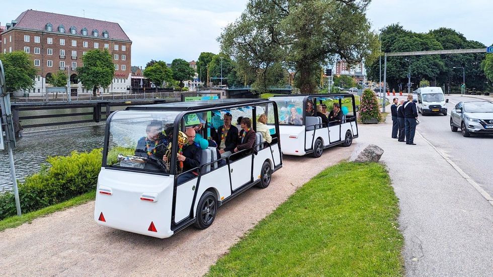 Electric trolleys on a riverside path in an urban area, passengers visible inside.