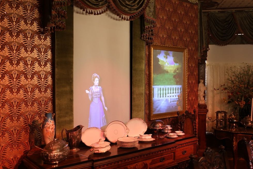 Elegant dining room with holographic woman, ornate wallpaper, and fine china display in Schifferdecker and Zelleken homes