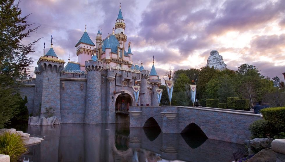 Enchanted castle at Disneyland with arched bridge at sunset, surrounded by trees and a cloudy sky.