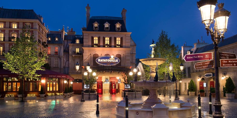 Evening view of Place de R\u00e9my at Disneyland Paris with fountain and lit street lamps.