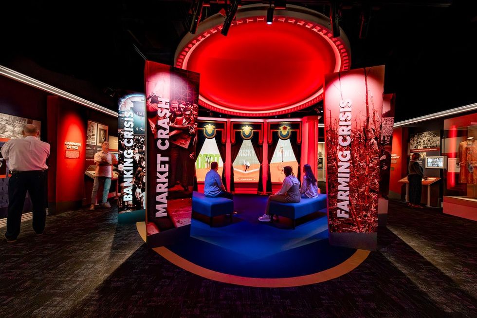 Exhibit at Herbert Hoover Presidential Library and Museum with red lighting showing banking, market, and farming crises. Visitors observe displays.