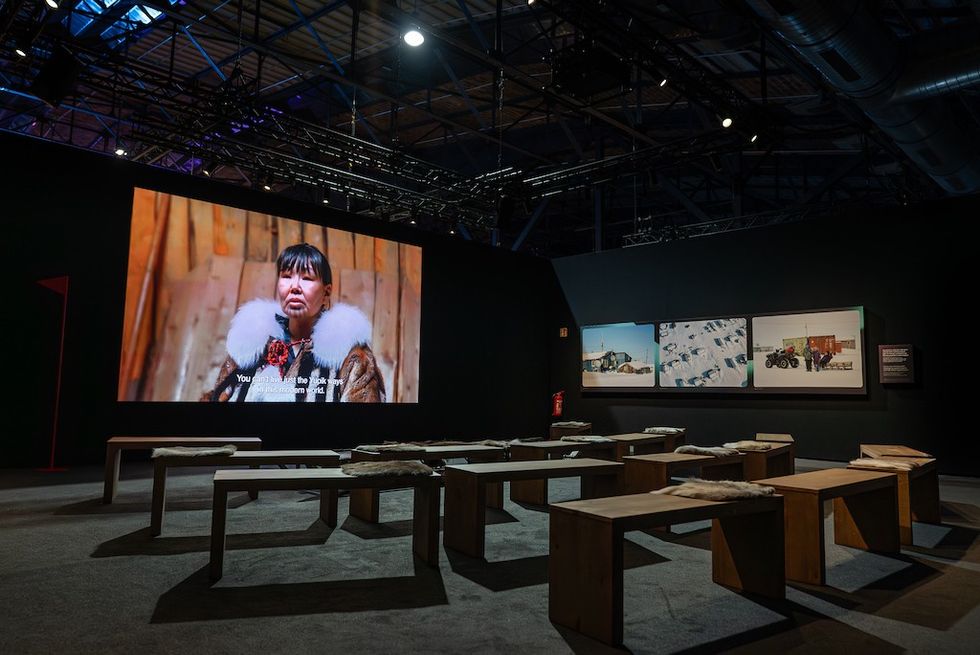 Exhibit with benches, large screen showing person, and framed photos on a dark wall.