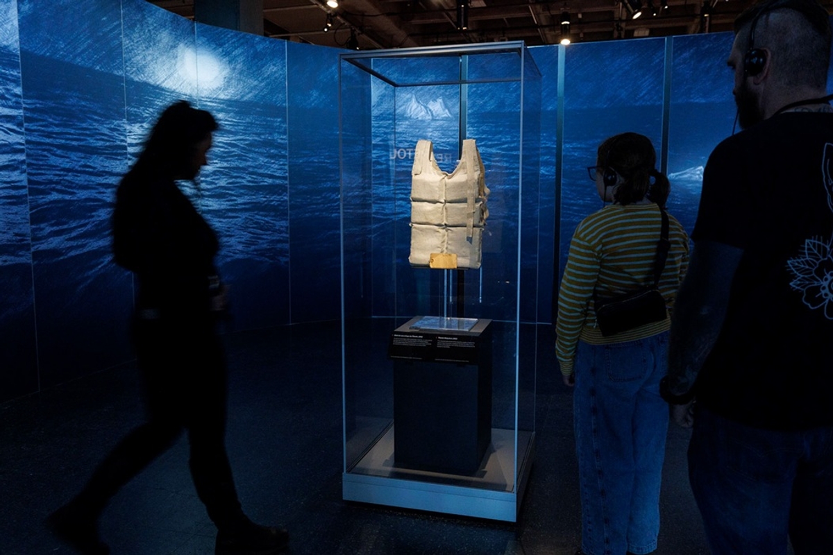 Exhibition visitors view a vintage life jacket in a glass case against a watery backdrop in Titanic: The Human Story