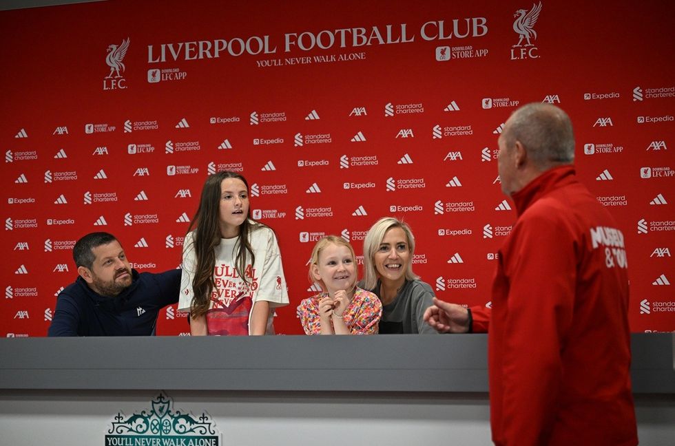 Family at Liverpool FC press room, smiling and talking with a staff member in red jacket.