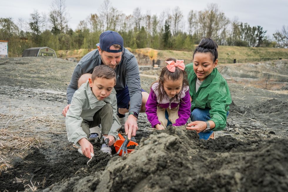 Family digging in dirt with tools at a park, surrounded by trees and open landscape.