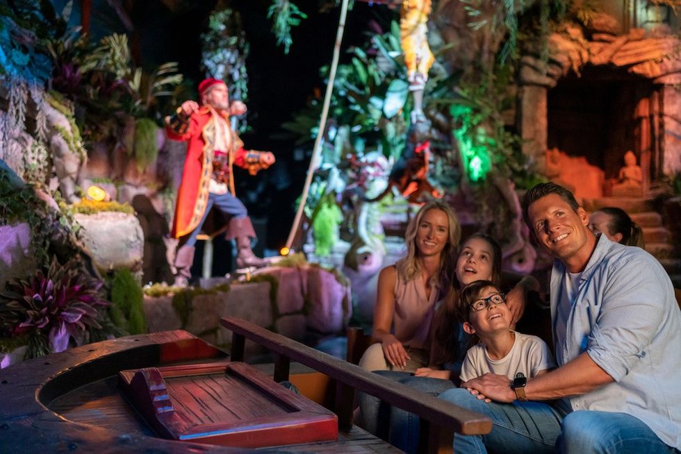 Family enjoying a pirate-themed boat ride with animatronic figure; lush jungle setting.