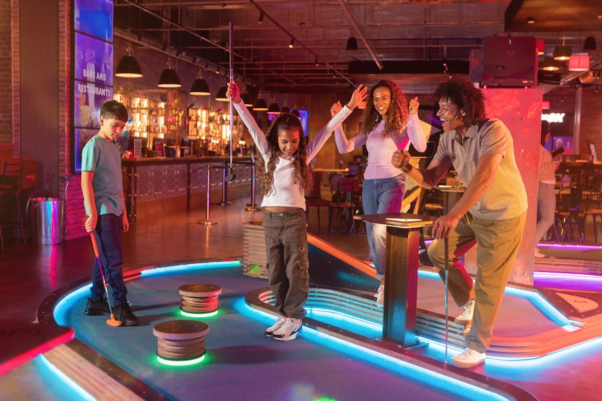Family enjoying mini-golf indoors with neon lights and a bar in the background.